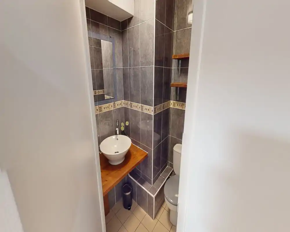 Bagno compatto e ben rifinito con pareti piastrellate grigie, lavabo a ciotola su piano in legno e wc; mensole e specchio offrono spazio funzionale.