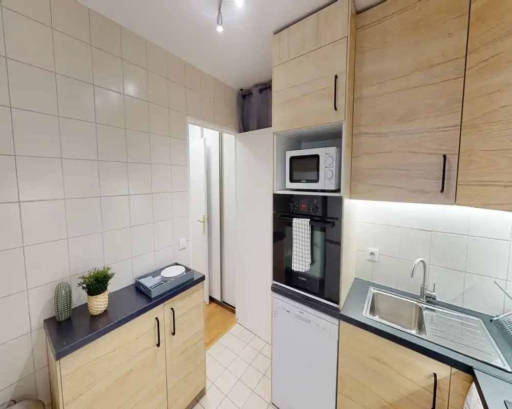 Cucina compatta e moderna con mobile effetto legno, forno e microonde incassati, lavastoviglie e lavabo in acciaio — ambiente luminoso e funzionale ideale per un piccolo appartamento.