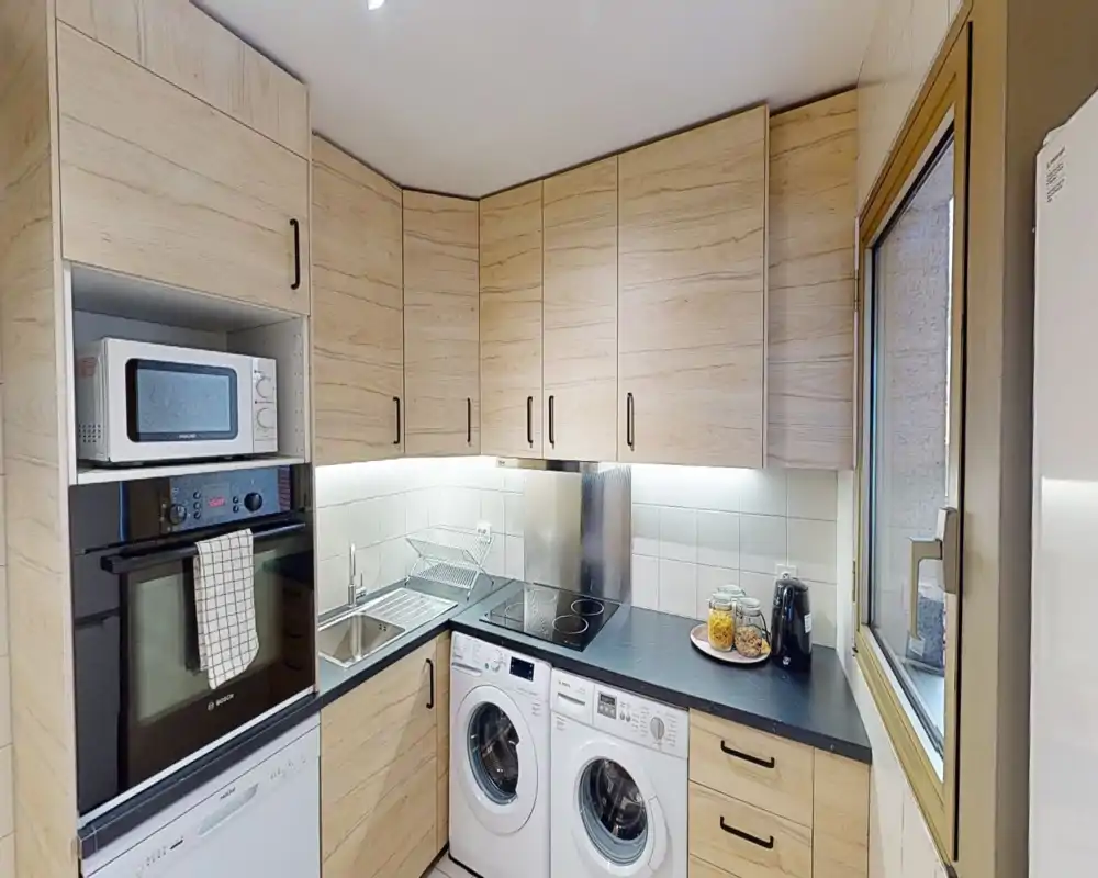 Cucina moderna compatta con mobili effetto legno, elettrodomestici integrati, illuminazione sotto i pensili e finestra che porta luce naturale — disposizione pratica con lavatrice e asciugatrice sotto il piano.