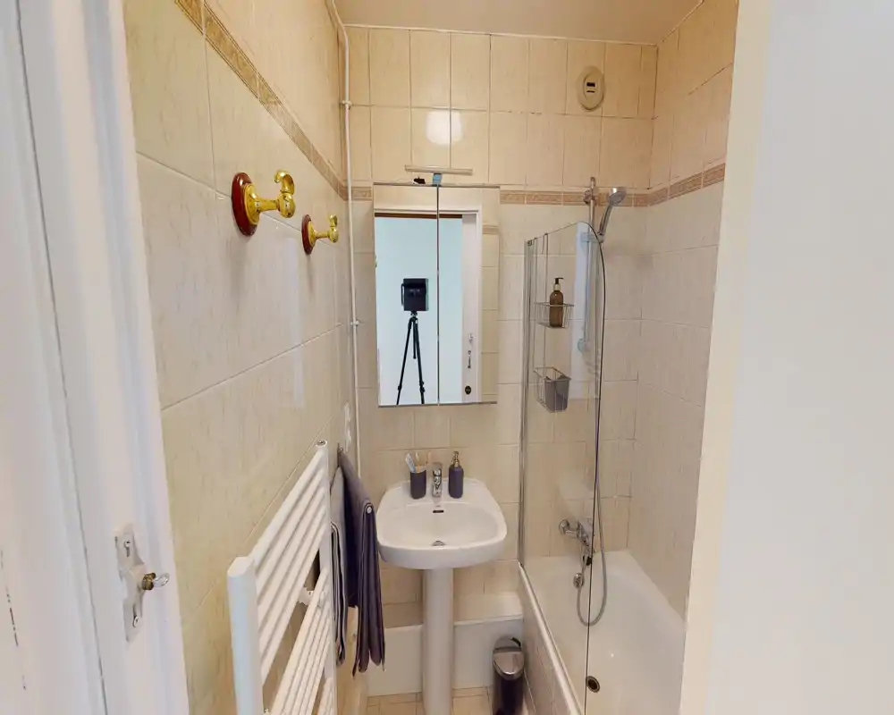 Bagno compatto e ben illuminato con lavabo a piedistallo, specchio con mobiletto, vasca con parete in vetro e pareti rivestite in piastrelle — ordinato e funzionale.