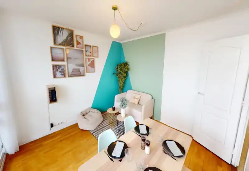 Coin salon lumineux et soigné avec un petit fauteuil, un pouf, un tapis et une table à manger au premier plan — décor moderne et chaleureux idéal pour un petit appartement.