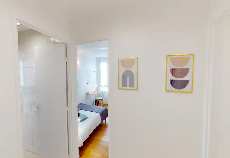 Chambre lumineuse et au style minimaliste vue depuis l'entrée. Lit double avec linge de lit blanc, jeté bleu foncé et un petit bureau près de la fenêtre, offrant un espace de nuit propre et fonctionnel.