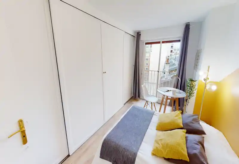 Chambre lumineuse et soignée avec lit double, coussins décoratifs et petite table près de la fenêtre-balcon — idéale pour voyageurs seuls ou couples.