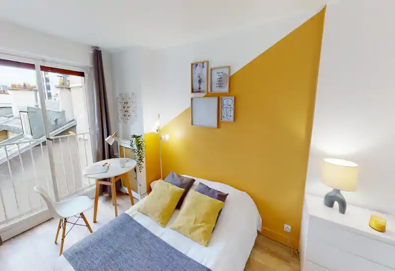 Chambre lumineuse et soignée avec un lit double devant un mur d'accent jaune moutarde, coussins décoratifs, lampe de chevet et une petite table près de la fenêtre apportant de la lumière naturelle.