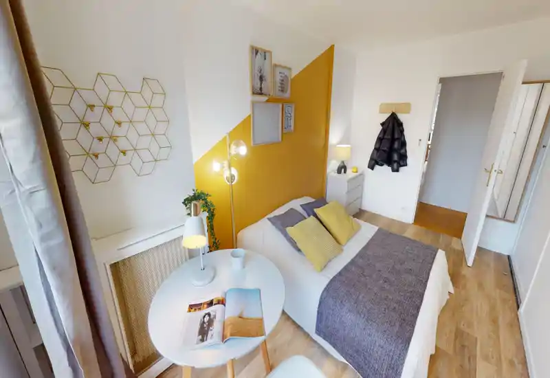 Chambre simple lumineuse et élégante avec un lit bien fait, un mur d'accent jaune et des textiles douillets. Une petite table ronde près de la fenêtre et un éclairage doux créent une atmosphère accueillante, parfaite pour la location courte durée.