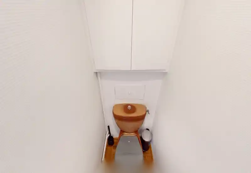 WC compact avec toilette, petite poubelle et brosse, murs blancs propres et sol en bois — adapté pour un petit sanitaire individuel.