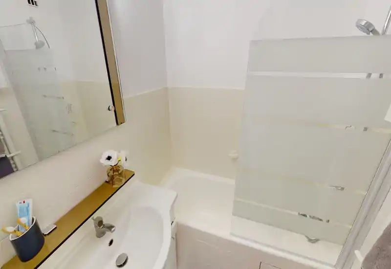 Salle de bains compacte et bien éclairée avec baignoire et pare-douche en verre, carrelage blanc et petit lavabo avec miroir — propre et fonctionnelle.