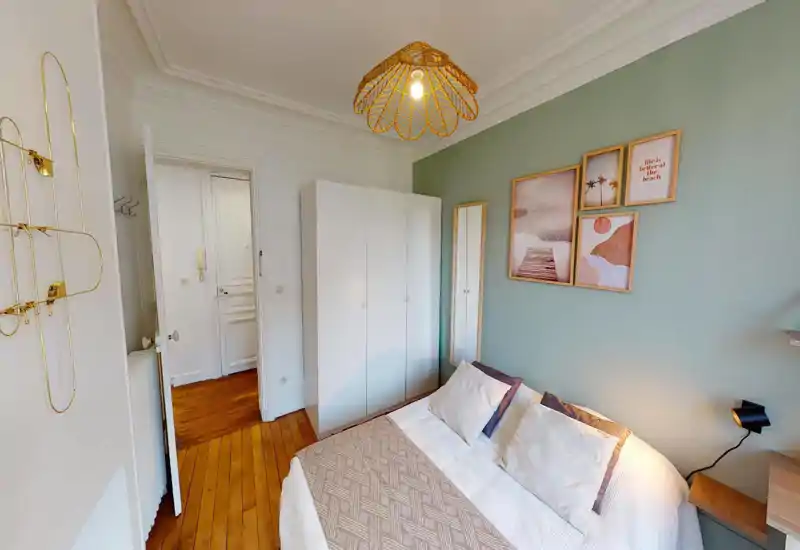 Bright, styled bedroom with a double bed dressed in neutral linens, decorative cushions and a textured throw. A white wardrobe and full-length mirror stand against a soft green accent wall adorned with framed artwork; warm wooden floors and a woven pendant lamp complete the cozy look.