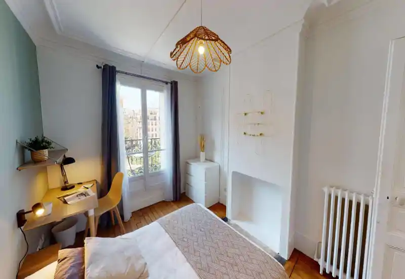 Bright, neatly styled bedroom with a large bed, wooden floors and a small workspace by the window. The room features soft textiles, a decorative pendant light and a dresser beside a decorative fireplace niche.