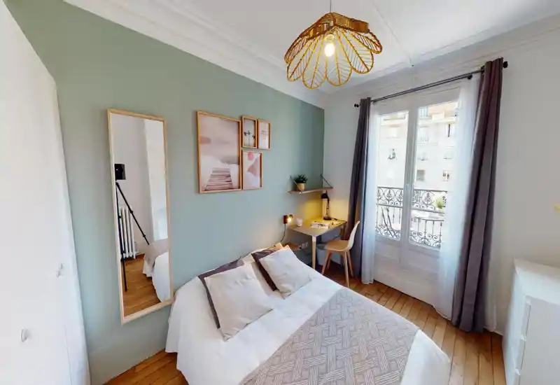 Bright, cozy bedroom with a double bed, decorative cushions and a soft throw. A small desk by the window and tasteful wall art create a pleasant, well-lit space ideal for relaxing or working.