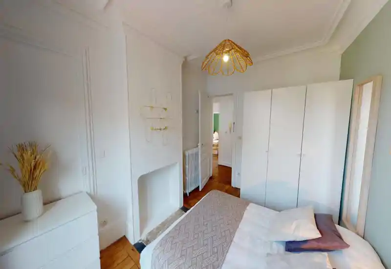 Bright, styled bedroom with a double bed, neutral linens and soft decorative pillows; white wardrobe and full-length mirror add storage and depth while warm wood floors and a woven pendant lamp create a cozy atmosphere.