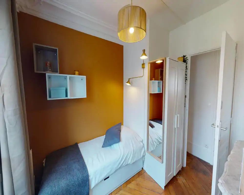 Camera singola compatta e accogliente con letto giorno ordinato, parete accentuata color ocra, mensole a muro e illuminazione soffusa — perfetta per un soggiorno breve.