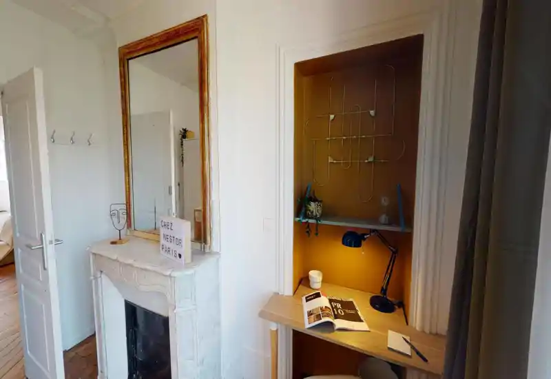 Angolo camera luminoso e stiloso con camino in marmo decorativo e un accogliente angolo studio con lampada e mensole; il letto è parzialmente visibile dalla porta aperta.