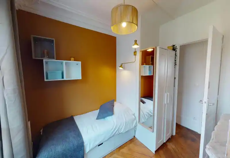 Camera singola compatta e accogliente con letto giorno ordinato, parete accentuata color ocra, mensole a muro e illuminazione soffusa — perfetta per un soggiorno breve.