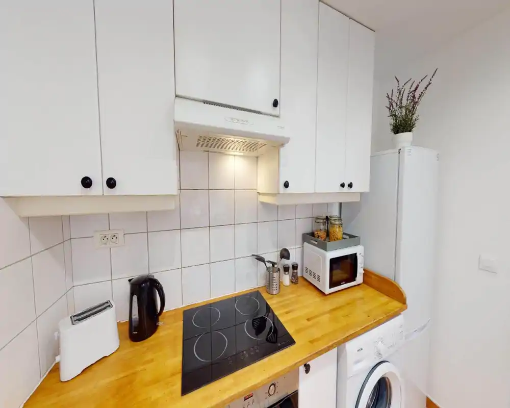 Cucina compatta e funzionale con piano in legno, piano a induzione, microonde, bollitore e frigorifero; pensili bianchi e rivestimento piastrellato donano un aspetto pulito e ordinato.