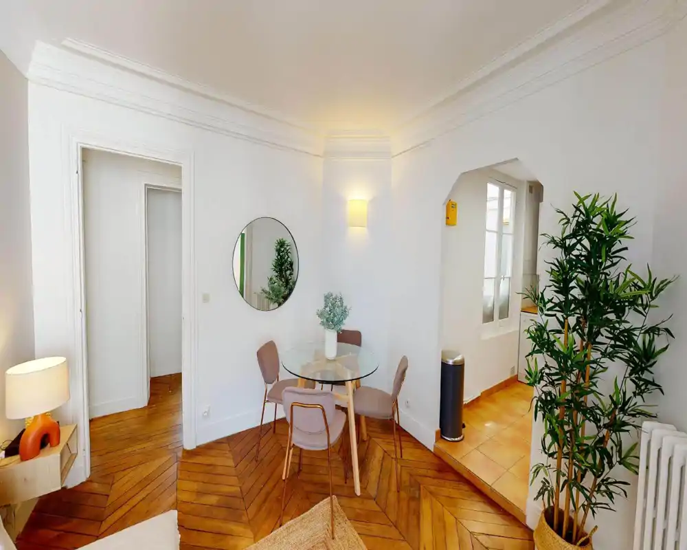 Accogliente angolo pranzo luminoso con tavolo rotondo in vetro, quattro sedie imbottite e pregiato parquet in legno. Decorazioni: specchio rotondo, piante e illuminazione calda per un'atmosfera intima.