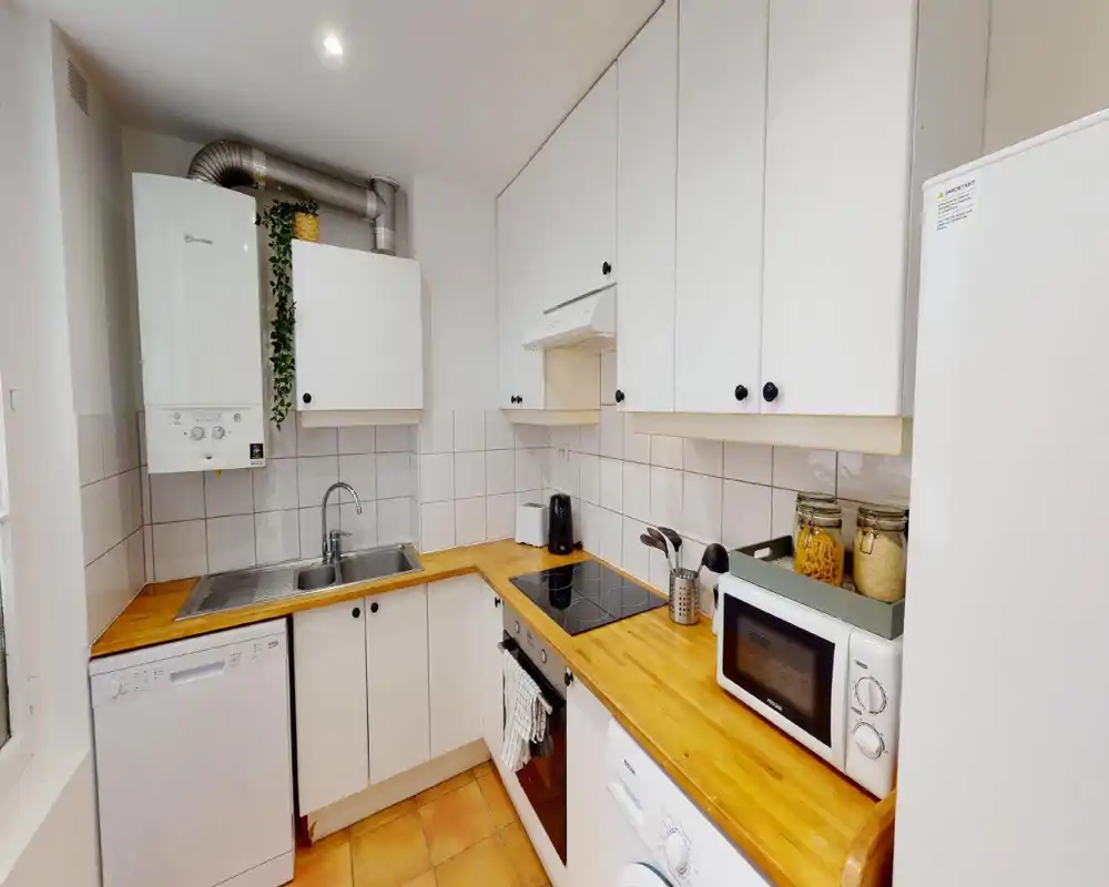 Cucina compatta e moderna con pensili bianchi, piani in legno, piano cottura in vetroceramica, forno integrato e frigorifero. Schienale piastrellato pulito, piano di lavoro ordinato con microonde e barattoli, atmosfera calda.