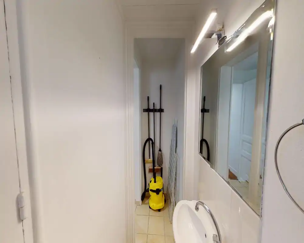 Piccolo bagno/area di servizio con lavabo, specchio e attrezzature per le pulizie riposte in una nicchia stretta. Piastrelle bianche neutre e disposizione pratica adatta a un bagno di servizio compatto.