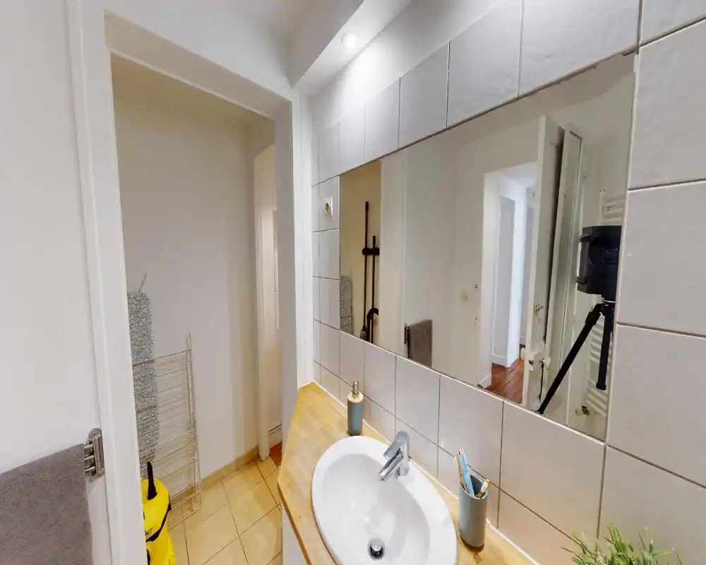 Area lavabo del bagno pulita e ben illuminata con grande specchio, piano in legno e piastrelle. Un portasciugamani, porta spazzolini e una piccola pianta aggiungono tocchi pratici.