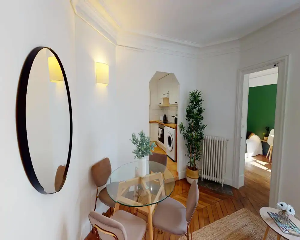 Zona pranzo luminosa e di stile con tavolo rotondo in vetro, quattro sedie imbottite e piante decorative — vista aperta su una cucina compatta e la camera adiacente.