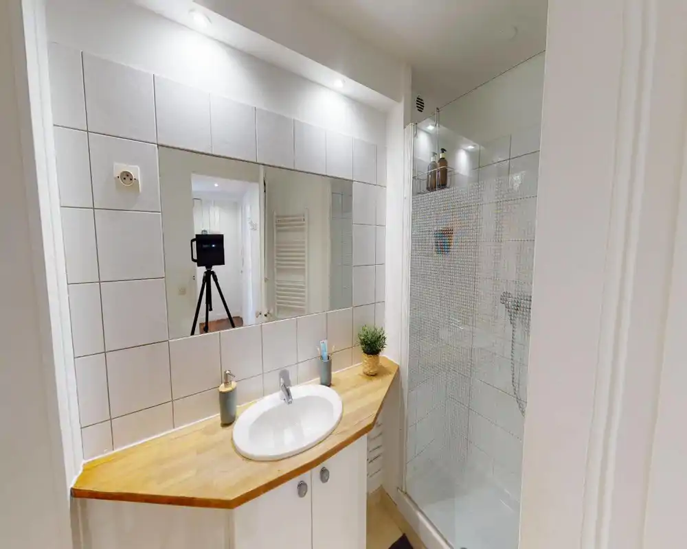 Bagno moderno e compatto con lavabo su piano in legno, grande specchio e doccia con parete in vetro. Piastrelle bianche e faretti incassati donano un aspetto pulito e luminoso.