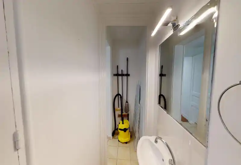 Small bathroom/utility area with a sink, mirror and cleaning equipment stored in a narrow alcove. Neutral white tiles and a practical layout suitable for a compact service bathroom.