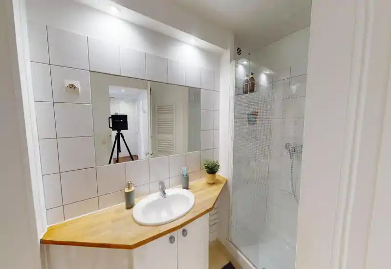 Modern, compact bathroom with a wooden countertop sink, large mirror, and a glass-enclosed shower. Bright white tiles and recessed lighting create a clean, fresh look.