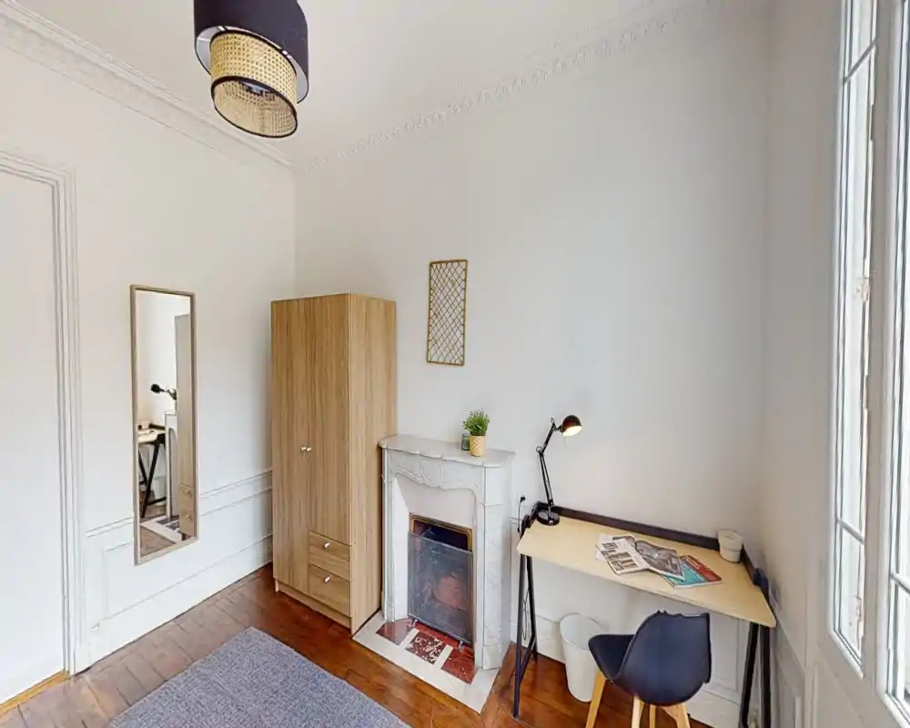 Compact bedroom-style room with a wardrobe, decorative fireplace and a simple workspace by a bright window. Light wooden floors and neutral walls create a calm, minimal atmosphere suitable for a single occupant.