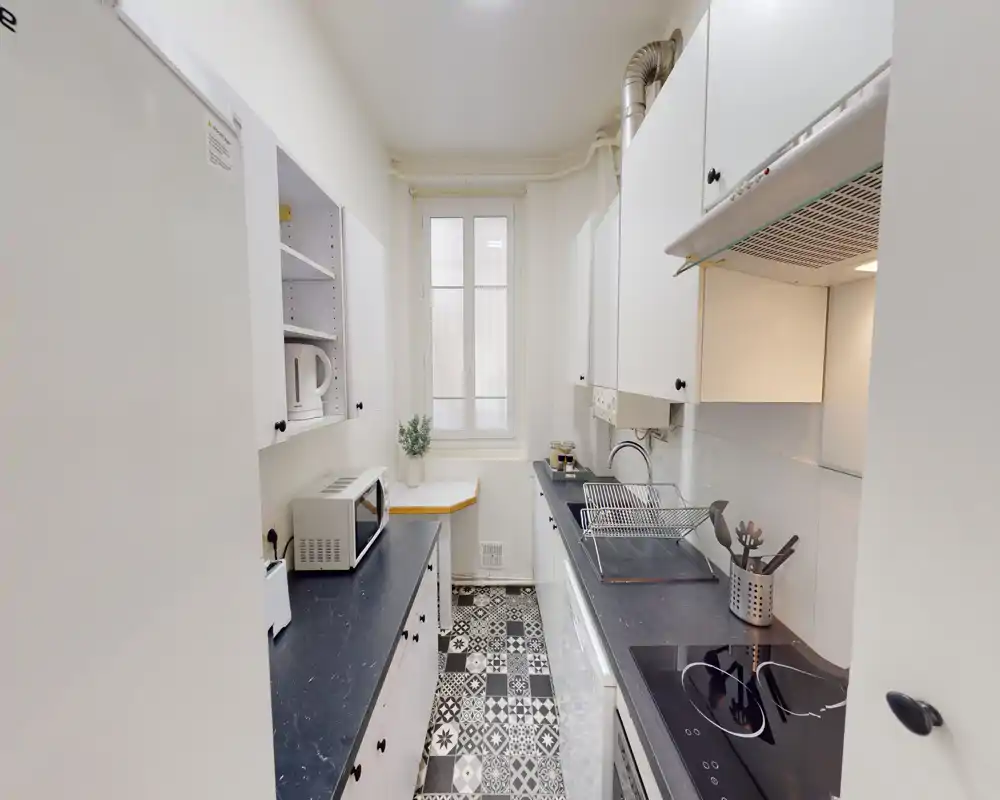 Bright, narrow kitchen with white cabinets, black countertops, patterned tile floor, electric cooktop and a sink — neat and well-equipped for a compact apartment.
