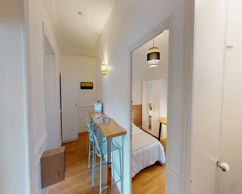 Compact, bright bedroom visible through a doorway with a white wardrobe and a double bed dressed in light linens. The adjacent hallway features a high counter and stools, offering a practical studio-like layout.