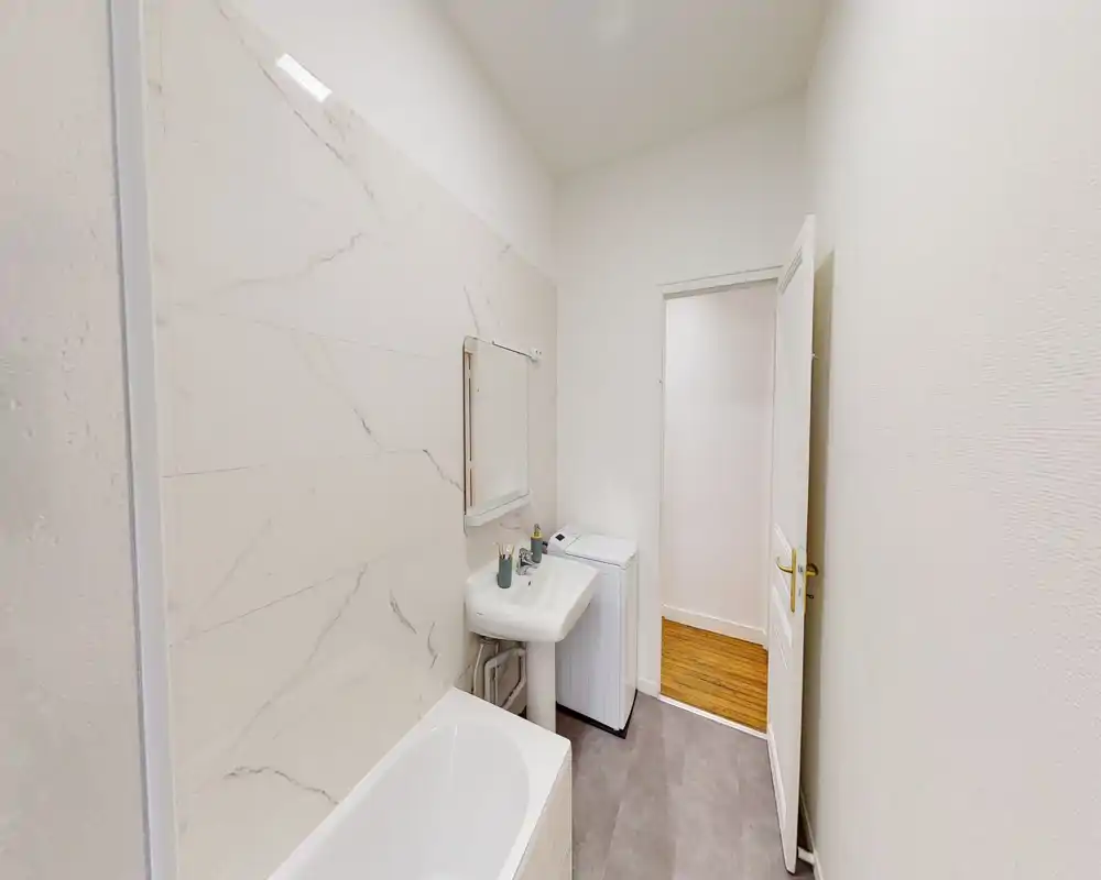 Compact, modern bathroom with a bathtub, wall-mounted sink and a washing machine. Neutral tiles and soft lighting create a clean, minimalist look ideal for rental listings.