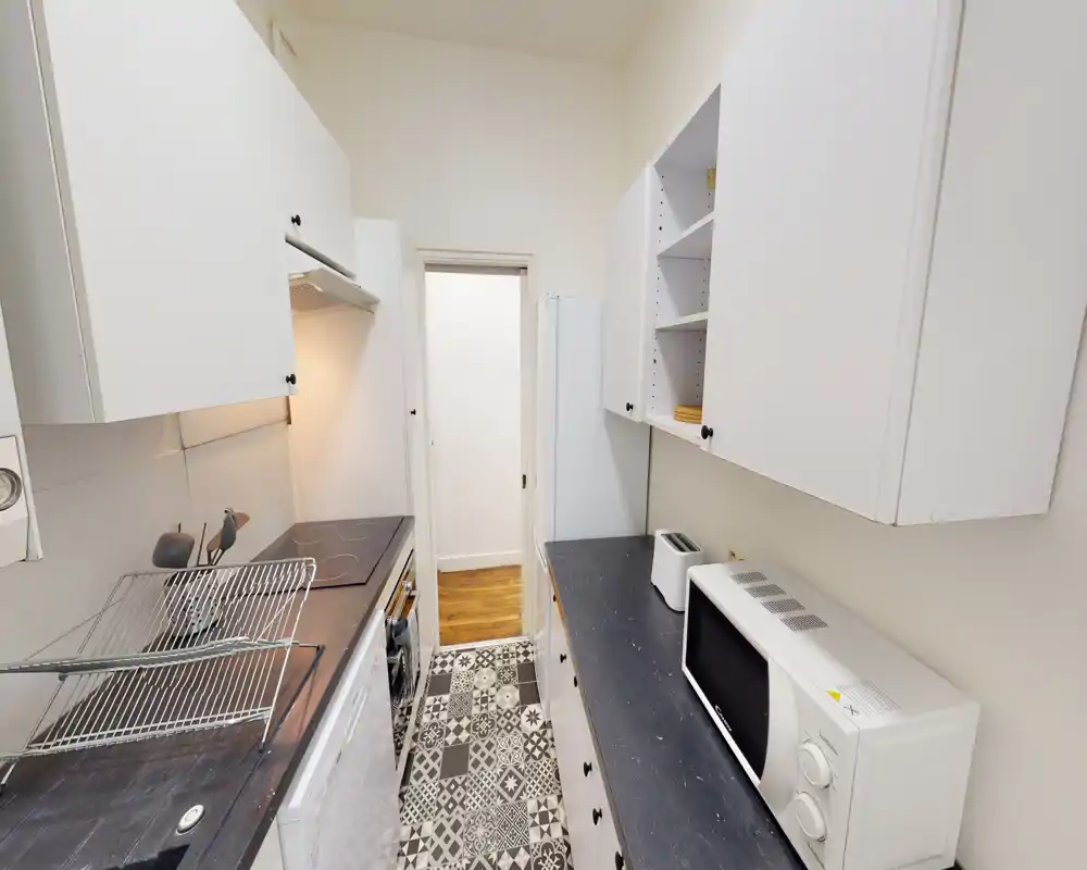 Compact galley kitchen with white cabinets, patterned tile floor and practical appliances including microwave and toaster — bright, tidy and efficiently laid out for city living.