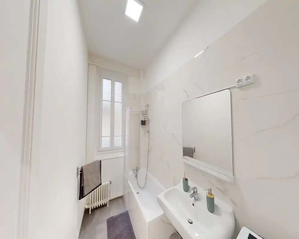 Clean, modern bathroom with a bathtub, wall-mounted showerhead, sink with mirror and subtle marble-effect wall tiles. Bright, neutral décor and practical towel and radiator near the window.
