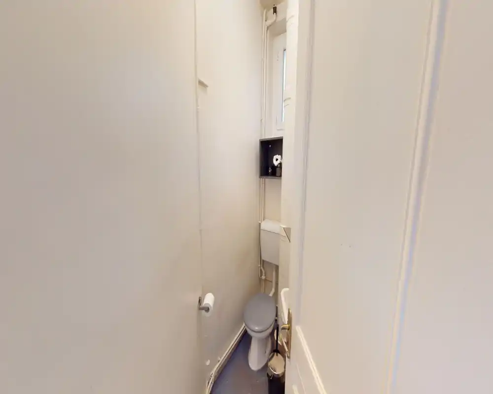 Compact single toilet room with a white WC, small cistern and shelving; narrow, clean space with neutral walls and natural light from a small high window.