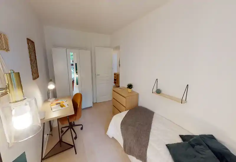 Camera da letto compatta e luminosa con letto singolo, comò, armadio con specchio e una piccola scrivania. Toni neutri e tessili morbidi creano un'atmosfera calma e accogliente, ideale per una persona o uno studente.