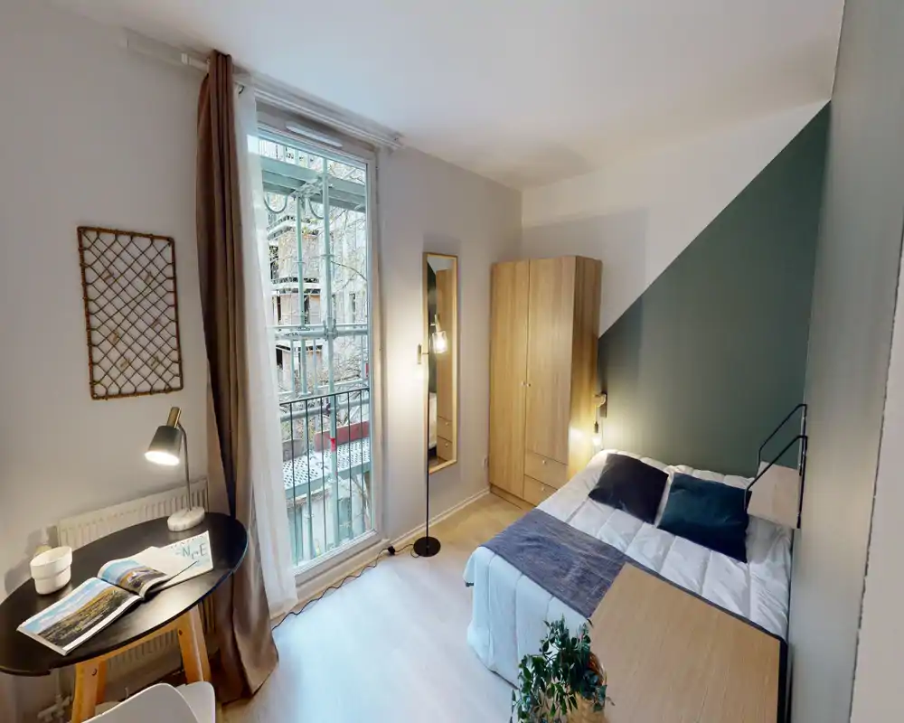 Chambre moderne et lumineuse avec un lit double confortable, des coussins décoratifs et une armoire en bois. Grande fenêtre apportant la lumière naturelle et un petit bureau formant un coin travail cosy.