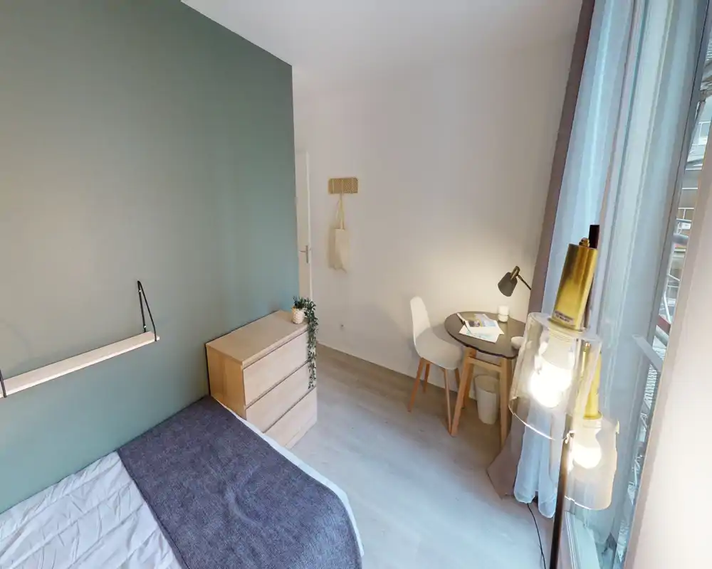 Chambre lumineuse et bien décorée, lit partiellement visible, commode et petit bureau près de la fenêtre — atmosphère chaleureuse.
