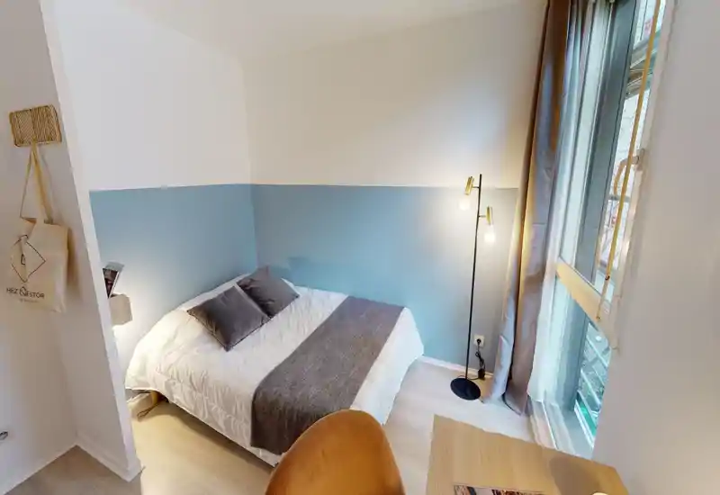 Sunlit, neatly styled small bedroom with a double bed dressed in white linens, decorative pillows and a gray runner; large window provides abundant natural light and a modern standing lamp adds warmth.