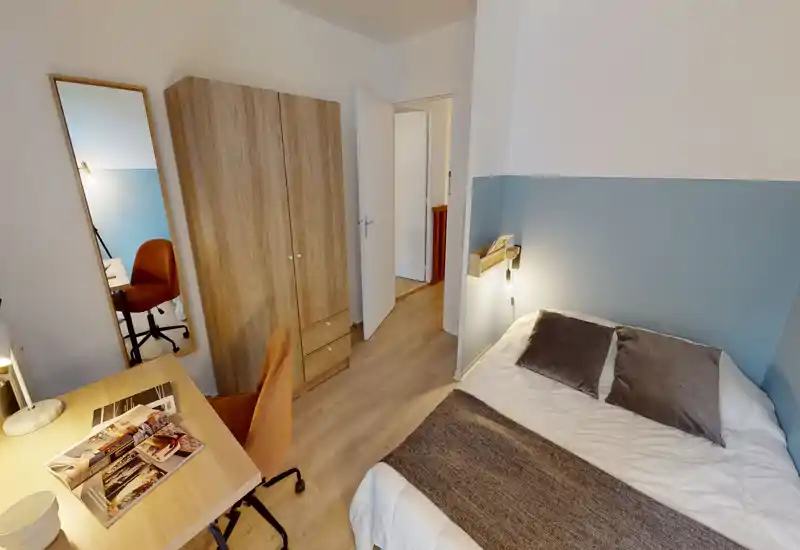 Cozy, well-staged single bedroom with a neatly made bed, bedside lighting and a wooden wardrobe. A compact desk and chair by the window create a practical workspace.