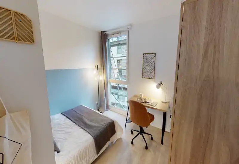 Bright, compact bedroom with a neatly made double bed, a study desk by the window and warm bedside lighting—ideal for a student or professional.