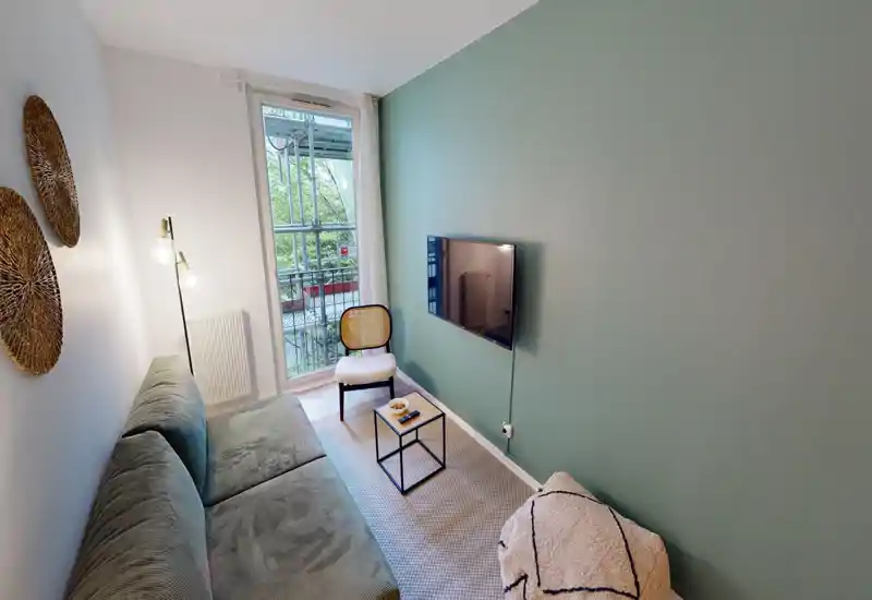 Bright, well-staged living room with a comfortable sofa, accent chair and wall-mounted TV facing a small coffee table; natural light from the balcony door enhances the calm, modern décor.