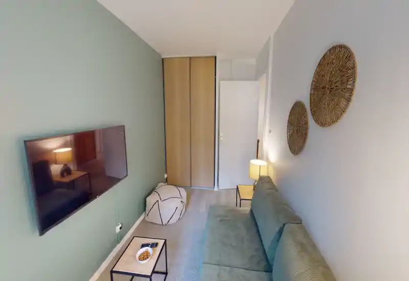 Soggiorno compatto con divano verde, TV a parete, decorazioni murali intrecciate e pouf — illuminazione calda e stile accogliente.