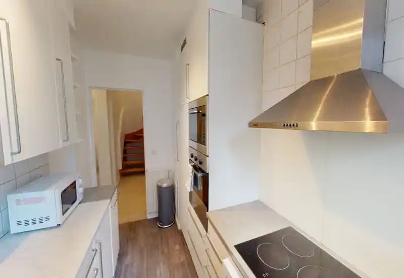 Narrow modern galley kitchen with white cabinets, stainless-steel hood and built-in ovens; clean worktops and an induction hob, ideal for compact apartments.