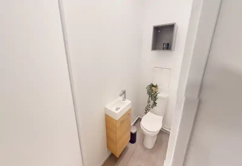 Compact, well-lit bathroom with a wall-mounted sink, modern toilet and small shelving — bright white walls and wood-look floor create a clean, minimal space.