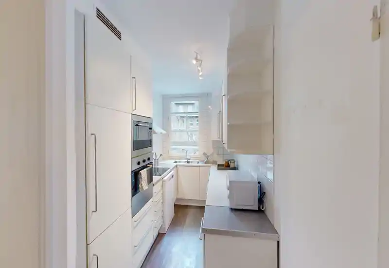 Bright, narrow galley kitchen with integrated appliances, white cabinets and a window at the far end letting in natural light.