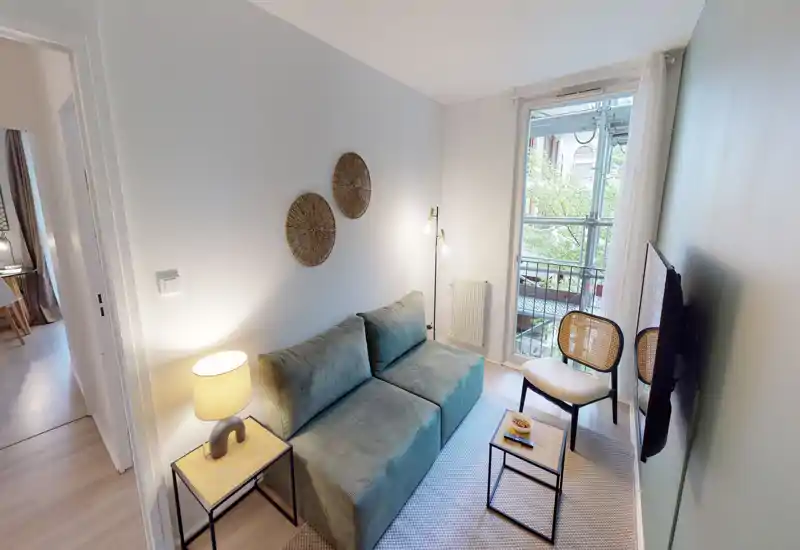 Soggiorno compatto e luminoso con divano a due posti, tavolino laterale con lampada, piccolo tavolino da caffè e sedie di fronte a una TV a parete. Grande finestra/porta si apre su un balcone, garantendo luce naturale e una vista piacevole.