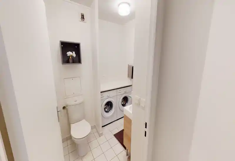 Compact bathroom with integrated laundry: toilet, sink and two front-loading washing machines under a countertop. Bright, clean white tiles and a simple modern style ideal for functional living.