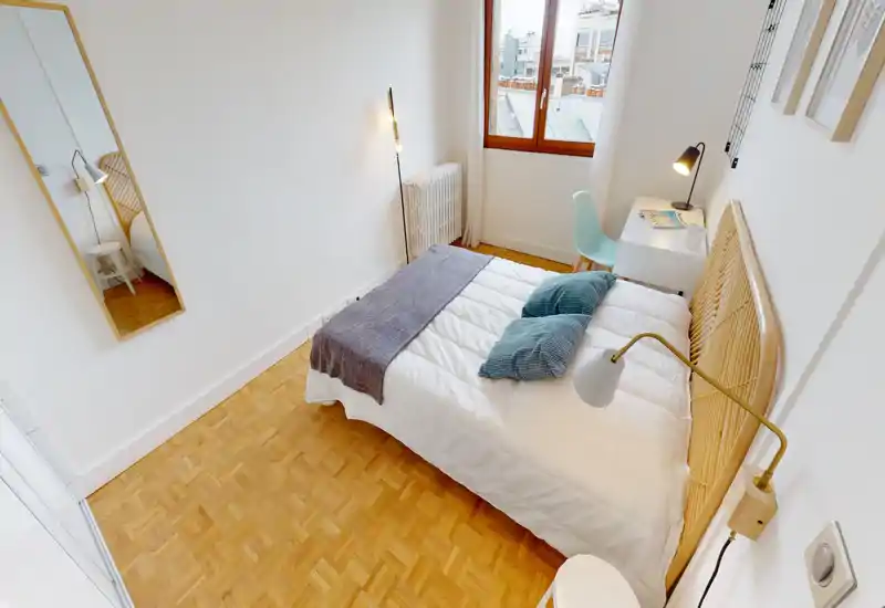 Chambre simple lumineuse et moderne avec tête de lit en rotin, literie blanche soignée, coussins décoratifs et un petit bureau près de la fenêtre — parfaite pour un séjour confortable en ville.
