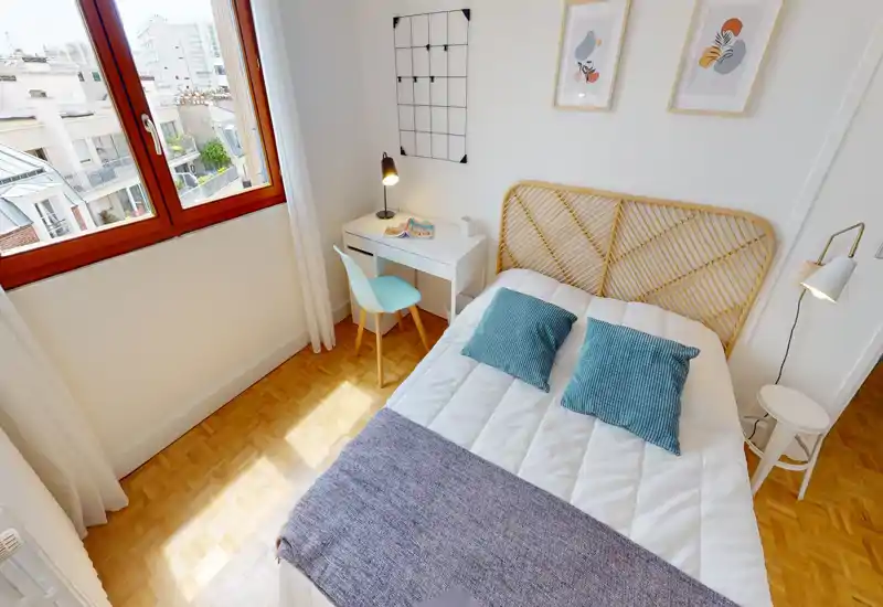 Chambre lumineuse et accueillante avec une tête de lit en rotin, deux coussins turquoise et un petit bureau blanc près d'une grande fenêtre laissant entrer beaucoup de lumière.