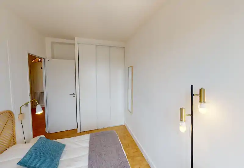 Chambre lumineuse et minimaliste avec lit partiellement visible, lampe de chevet et lampadaire. Murs blancs et parquet pour une atmosphère propre et aérée.